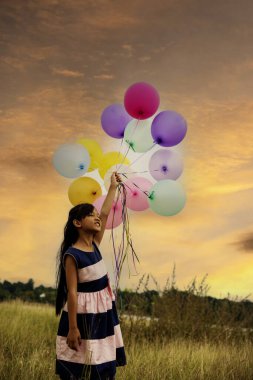 Dikey gün doğumu altın renkli gökyüzü elleri balonları tutuyor. Yeşil çayır mutluluğuyla koşan neşeli tatlı kız. Hava balonları tutan eller doğum günü partisinde dışarıda mutlu bir altın gökyüzü oynar.