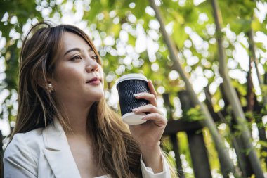 Yeşil Park 'ta kahve içen bir kadın elinde sıcak tek kullanımlık bardak tutuyor. Happy rahatla güzel Asyalı kadın gülümseyen yüz açık hava bahçesinde duruyor. Genç kadınlar doğanın tadını çıkarıyorlar Özgürlük yaşam tarzı.