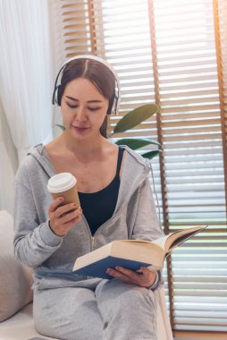 Mutlu Asyalı kadın kulaklık takıyor, kitap okuyor, kahve içiyor evdeki oturma odasında rahat bir kanepede oturuyor. Kadın Asyalılar evde çalışır, kitap okurlar, mutlu okuyucular gibi yaşarlar.