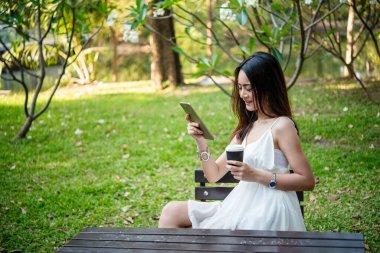 Asyalı kadın yeşil parkta tek kullanımlık sıcak kahve bardağı tutarken akıllı telefon kullanıyor. Banner Happy rahat Asyalı kadın açık hava bahçesinde gülümsüyor. Genç kadınlar fotokopi odasıyla doğanın tadını çıkarıyor