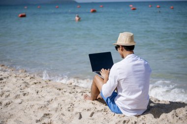 Erkek gezgin yaz aylarında mavi gökyüzünde dizüstü bilgisayar kullanır. Asyalı adam tropikal gömlek tarzında günlük pantalon giyer yazın plajda dinlenir. Dizüstü bilgisayarda yazı yazan adam tatil tatili gezisi