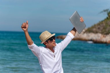 Erkek gezgin yaz aylarında mavi gökyüzünde dizüstü bilgisayar kullanır. Asyalı adam tropikal gömlek tarzında günlük pantalon giyer yazın plajda dinlenir. Dizüstü bilgisayarda yazı yazan adam tatil tatili gezisi