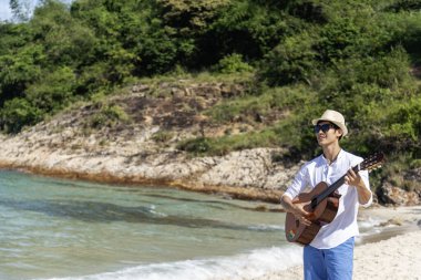 Genç erkekler plajda akustik gitar çalıyorlar. Akustik gitar enstrümanı çalan mutlu bir adam kumsalda, tropik plaj adasında oturuyor. Asyalı müzisyen Mutlu gitarist hobi yaşam tarzı