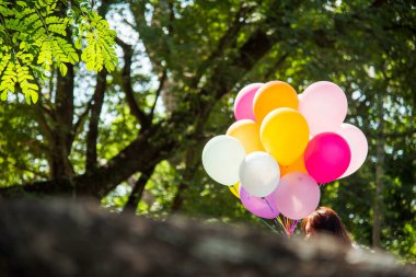 Balon tutan neşeli güzel kadın yeşil parktaki büyük ağacın altında mutlu mesut oturuyor. Kadın Eller 'in elinde canlı hava balonları var. Doğum günü partisinde oynuyorlar. Mutlu yaz mevsimi açık havada.