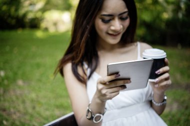 Asyalı kadın yeşil parkta tek kullanımlık sıcak kahve bardağı tutarken akıllı telefon kullanıyor. Banner Happy rahat Asyalı kadın açık hava bahçesinde gülümsüyor. Genç kadınlar fotokopi odasıyla doğanın tadını çıkarıyor