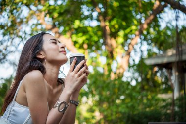 Yeşil Park 'ta kahve içen bir kadın elinde sıcak tek kullanımlık bardak tutuyor. Happy rahatla güzel Asyalı kadın gülümseyen yüz açık hava bahçesinde duruyor. Genç kadınlar doğanın tadını çıkarıyorlar Özgürlük yaşam tarzı.