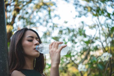 Su içen genç bir kadın açık hava parkı yolcusu. Asyalı kadın içme suyu şişesi sağlıklı insan mutlu bir gülümseme. Mutluluk. Güzel Asyalı Çinli kadınlar ellerinde maden suyu şişesi yaşam tarzı tutuyorlar.