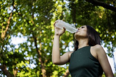 Su içen genç bir kadın açık hava parkı yolcusu. Asyalı kadın içme suyu şişesi sağlıklı insan mutlu bir gülümseme. Mutluluk. Güzel Asyalı Çinli kadınlar ellerinde maden suyu şişesi yaşam tarzı tutuyorlar.