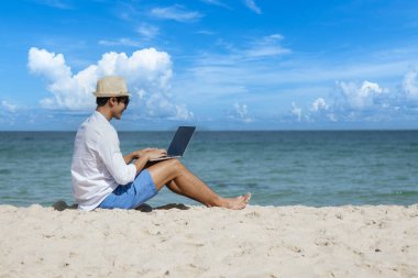 Erkek gezgin yaz aylarında mavi gökyüzünde dizüstü bilgisayar kullanır. Asyalı adam günlük mavi şort beyaz gömlek yaz plajında oturarak rahatlıyor. Adada dizüstü bilgisayarda yazı yazan adam tatil tatili iş seyahatinde serbest çalışıyor.