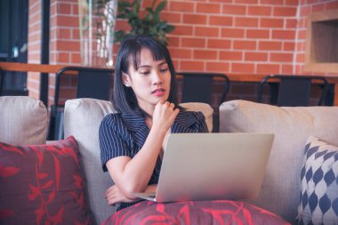 Mutlu Asyalı kadın video konferansı evdeki oturma odasında rahat koltukta oturuyor. Gülümseyen Asyalı kadın dizüstü bilgisayar kulaklığı kullanıyor. Evlerinden video izliyorlar.