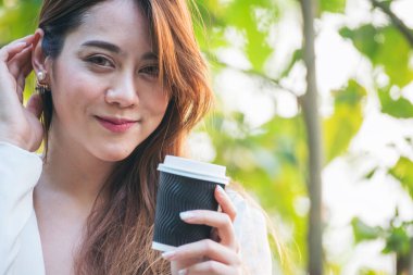 Yeşil Park 'ta kahve içen bir kadın elinde sıcak tek kullanımlık bardak tutuyor. Happy rahatla güzel Asyalı kadın gülümseyen yüz açık hava bahçesinde duruyor. Genç kadınlar doğanın tadını çıkarıyorlar Özgürlük yaşam tarzı.