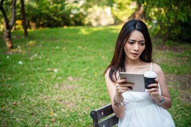 Asyalı kadın yeşil parkta tek kullanımlık sıcak kahve bardağı tutarken akıllı telefon kullanıyor. Banner Happy rahat Asyalı kadın açık hava bahçesinde gülümsüyor. Genç kadınlar fotokopi odasıyla doğanın tadını çıkarıyor