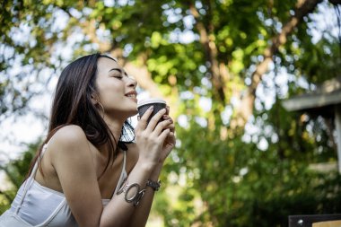 Yeşil Park 'ta kahve içen bir kadın elinde sıcak tek kullanımlık bardak tutuyor. Happy rahatla güzel Asyalı kadın gülümseyen yüz açık hava bahçesinde duruyor. Genç kadınlar doğanın tadını çıkarıyorlar Özgürlük yaşam tarzı.