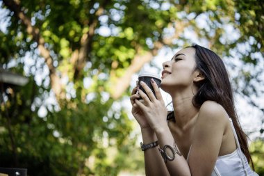 Yeşil Park 'ta kahve içen bir kadın elinde sıcak tek kullanımlık bardak tutuyor. Happy rahatla güzel Asyalı kadın gülümseyen yüz açık hava bahçesinde duruyor. Genç kadınlar doğanın tadını çıkarıyorlar Özgürlük yaşam tarzı.