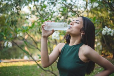 Su içen genç bir kadın açık hava parkı yolcusu. Asyalı kadın içme suyu şişesi sağlıklı insan mutlu bir gülümseme. Mutluluk. Güzel Asyalı Çinli kadınlar ellerinde maden suyu şişesi yaşam tarzı tutuyorlar.