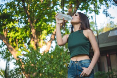 Su içen genç bir kadın açık hava parkı yolcusu. Asyalı kadın içme suyu şişesi sağlıklı insan mutlu bir gülümseme. Mutluluk. Güzel Asyalı Çinli kadınlar ellerinde maden suyu şişesi yaşam tarzı tutuyorlar.