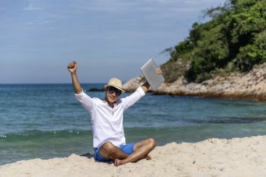 Erkek gezgin yaz aylarında mavi gökyüzünde dizüstü bilgisayar kullanır. Asyalı adam tropikal gömlek tarzında günlük pantalon giyer yazın plajda dinlenir. Dizüstü bilgisayarda yazı yazan adam tatil tatili gezisi