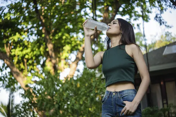 Su içen genç bir kadın açık hava parkı yolcusu. Asyalı kadın içme suyu şişesi sağlıklı insan mutlu bir gülümseme. Mutluluk. Güzel Asyalı Çinli kadınlar ellerinde maden suyu şişesi yaşam tarzı tutuyorlar.