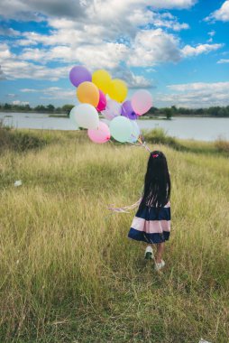 Dikey neşeli kız elinde balonlarla yeşil çayır üzerinde koşuyor mutlulukla mavi bulutlu gökyüzü. Doğum günü partisinde hava balonları tutan eller dışarıda mutlu zamanlar geçirir.