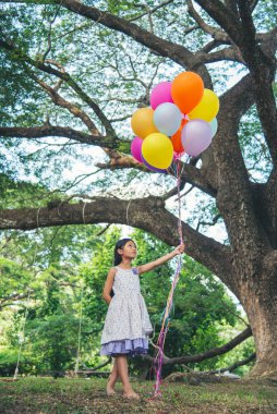 Dikey Neşeli Güzel Kadın elinde balonlarla yeşil parktaki büyük ağacın altında mutlu mesut oturuyor. Kadın Eller 'in elinde canlı hava balonları var. Yaz tatilinde dışarıda doğum günü partisi yapıyorlar.