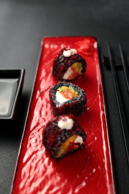Mürekkepbalığı mürekkebi, mango, somon, tobiko havyarı ve avokado içeren suşi rulosu siyah arka planda sergilenir. Japon mutfağının renkli ve lezzetli bir temsili, stok fotoğrafçılığı için mükemmel..