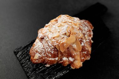 Bademli kruvasan. Puf böreği, yağla zenginleştirilmiş bademli narin frangipan kreması. En üstte, kruvasanın biraz badem gevreği ve pudra şekeri var. Siyah bir kesme tahtasının üzerinde