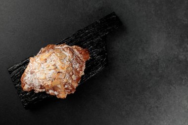 Bademli kruvasan. Puf böreği, yağla zenginleştirilmiş bademli narin frangipan kreması. En üstte, kruvasanın biraz badem gevreği ve pudra şekeri var. Siyah bir kesme tahtasının üzerinde