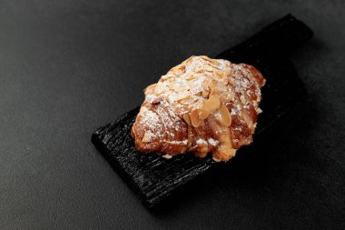 Bademli kruvasan. Puf böreği, yağla zenginleştirilmiş bademli narin frangipan kreması. En üstte, kruvasanın biraz badem gevreği ve pudra şekeri var. Siyah bir kesme tahtasının üzerinde