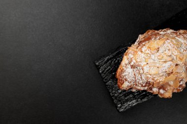 Bademli kruvasan. Puf böreği, yağla zenginleştirilmiş bademli narin frangipan kreması. En üstte, kruvasanın biraz badem gevreği ve pudra şekeri var. Siyah bir kesme tahtasının üzerinde