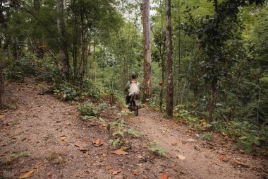 Ormanda elinde tüfekle motosiklet süren bir adam. Pai, Kuzey Tayland.