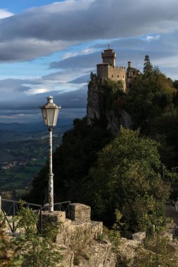 San Marino 'da kayalık bir uçurumun tepesinde tarihi bir kule ve ön planda klasik sokak lambası var.