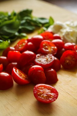 Arka planda fesleğen yaprakları ve mozzarella olan ahşap bir tahtada taze kiraz domatesleri..