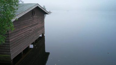 Sisli bir sabahta ağaçtan bir kayıkhane ve yanından geçen bir motorlu tekne..