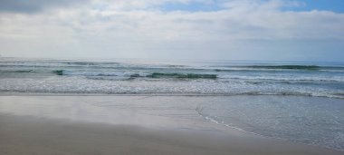 Güneşli bir günde kumsalın beyaz kumlu ve sakin denizi sahile vuran yüzücüler ve sörfçülerle.