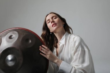 Güzel genç bir kadın, beyaz duvarda asılı davulla rahatlatıcı müzik çalar. Yüksek kalite fotoğraf.