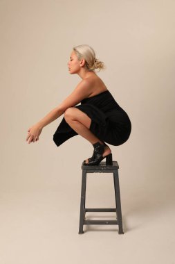 Beyaz duvarın üzerindeki taburede oturan siyah elbiseli güzel sarışın genç kadın. Yüksek kalite fotoğraf.