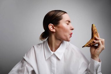 Beyaz gömlek giyen, elinde olgun bir muz tutan ve suratını buruşturan genç bir kadının portresi. Yüksek kalite fotoğraf.