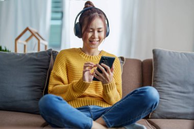 Kanepede otururken kulaklık takan Asyalı genç bir kadının portresi..