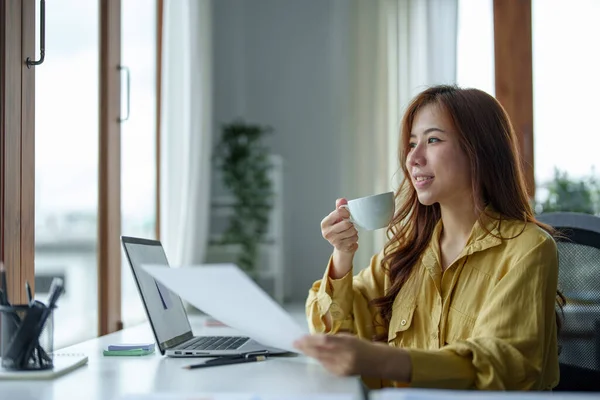 一个快乐的亚洲女人在日间喝咖啡休息时坐在办公桌前微笑的画像 — 图库照片