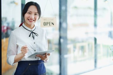 Küçük bir işletme kuran ve açan genç bir Asyalı kadın, elinde tablet tutan gülümseyen bir yüz ile bir kahve dükkanının önünde duruyor. İş sahibi, restoran, kafe konsepti.