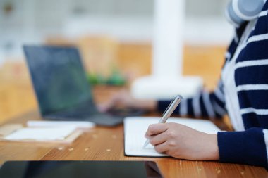 Bilgisayar kullanan genç Asyalı bir kadının portresi, kulaklık takıyor ve kütüphanedeki ahşap bir masada video konferans yoluyla ders çalışmak için bir not defteri kullanıyor..