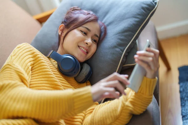 Portret Uśmiechniętej Azjatki Noszącej Słuchawki Używającej Telefonu Słuchającej Muzyki Siedzącej — Zdjęcie stockowe