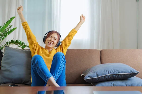 stock image Portrait of a smiling Asian woman wearing a pair of headphones and using her phone and listening to music while sitting on the sofa.