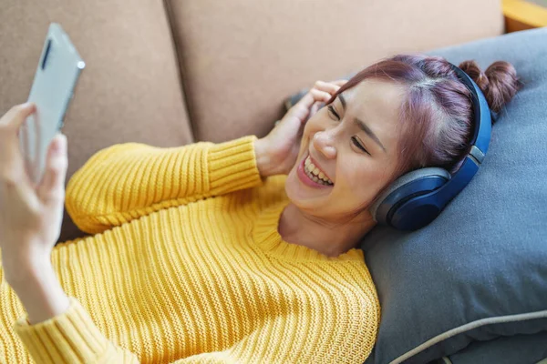 一个笑着的亚洲女人坐在沙发上 戴着耳机 用手机听音乐的画像 — 图库照片