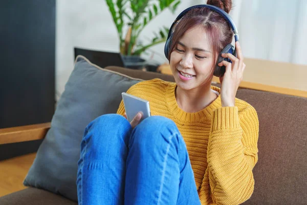 一个笑着的亚洲女人坐在沙发上 戴着耳机 用手机听音乐的画像 — 图库照片