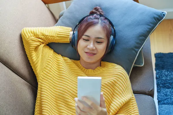 Portret Uśmiechniętej Azjatki Noszącej Słuchawki Używającej Telefonu Słuchającej Muzyki Siedzącej — Zdjęcie stockowe