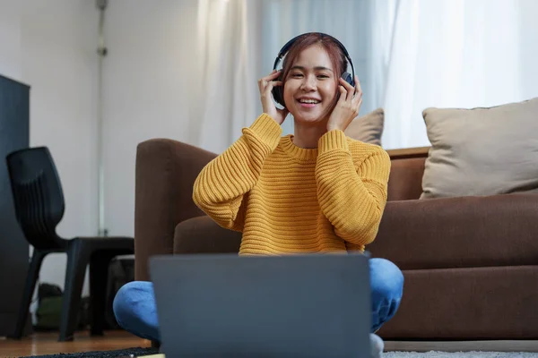 一个年轻的亚洲女人的画像 她面带微笑 头戴耳机 用电脑 坐在沙发上听音乐 — 图库照片