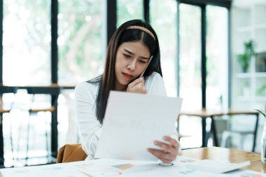 Masasındaki telefon ve mali belgeleri kullanırken stresli yüzünü gösteren güzel bir Asyalı kadın çalışanın portresi..