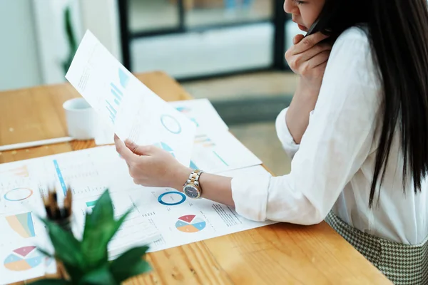 stock image A portrait of a beautiful Asian female employee showing a stressed face while using the phone and financial documents on her desk.