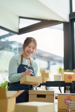 Küçük bir girişimin portresi, ve KME sahibi, Asyalı bir kadın girişimci, müşterinin iç kutusuna koymadan önce ürünü organize etmek için bir not defterine bilgi yazıyor..